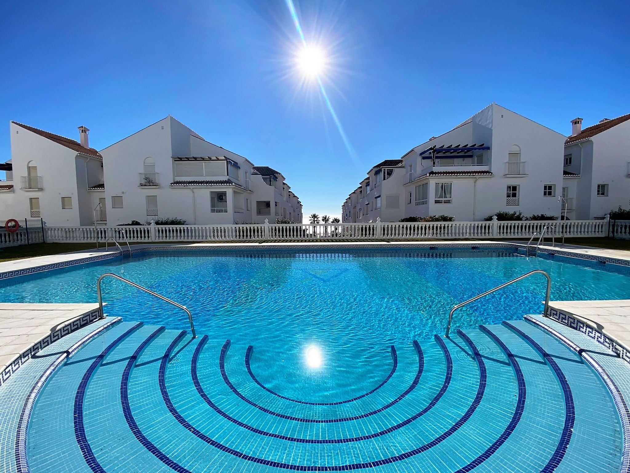 A swimming pool with clear blue water, surrounded by white buildings with balconies, under a clear sky.