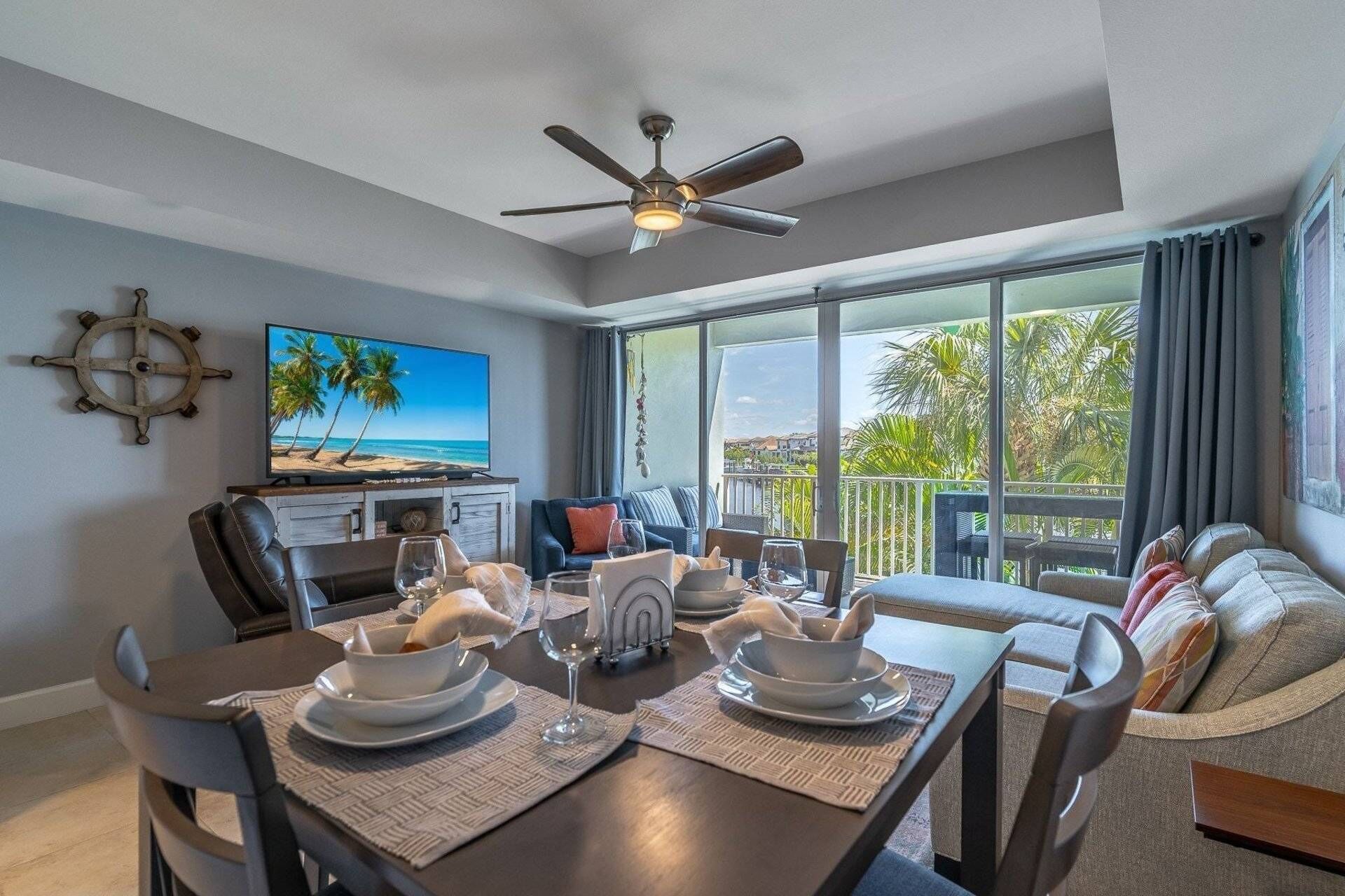 A modern living room with a dining area, a television displaying a beach scene, a ceiling fan, and a balcony with a view of palm trees.