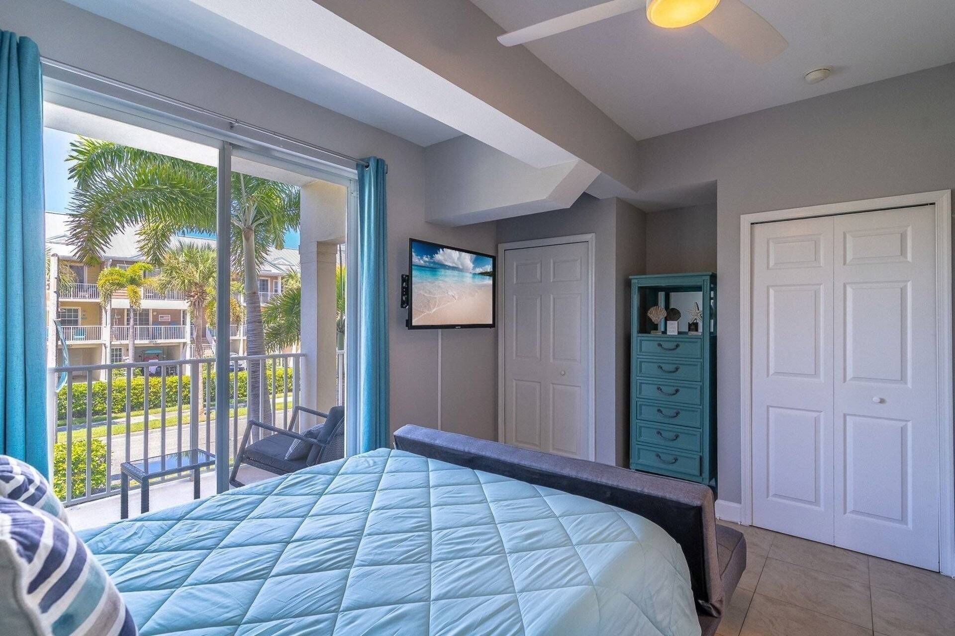 A hotel room with a bed, a television, a wardrobe, and a balcony with a view of palm trees and a building.