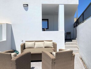 Uma área de estar moderna ao ar livre, com um sofá, cadeiras, uma mesa pequena e uma janela com vista para um edifício.