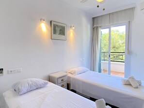 Habitación de hotel con dos camas individuales, una mesita de noche pequeña con cajones, un cuadro en la pared y una ventana con vistas a la vegetación.