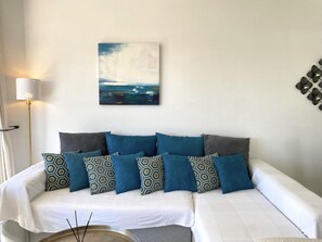 Ein Schlafzimmer mit einem weißen Bett, blau gemusterten Kissen, einem Bild an der Wand und einer Lampe.