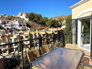 Ein Balkon mit Glastisch und Korbstühlen, der einen Blick auf eine Hügelside mit weißen Gebäuden und roten Ziegeldächern bietet.