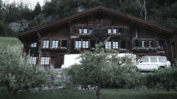 Un rustico chalet in legno con balcone, immerso nel verde e con un'auto parcheggiata.