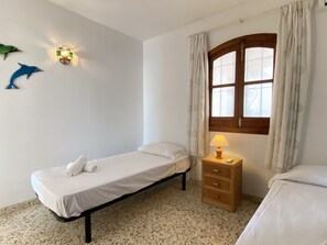 A small, simple room with a bed, a nightstand, a lamp, a window with curtains, and wall decorations including two fish. - Apartment  by Interhome (Nerja)