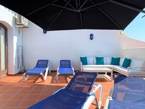 Eine Terrasse mit zwei Liegestühlen, einem Sofa und einem Tisch, beschattet durch einen großen Sonnenschirm.