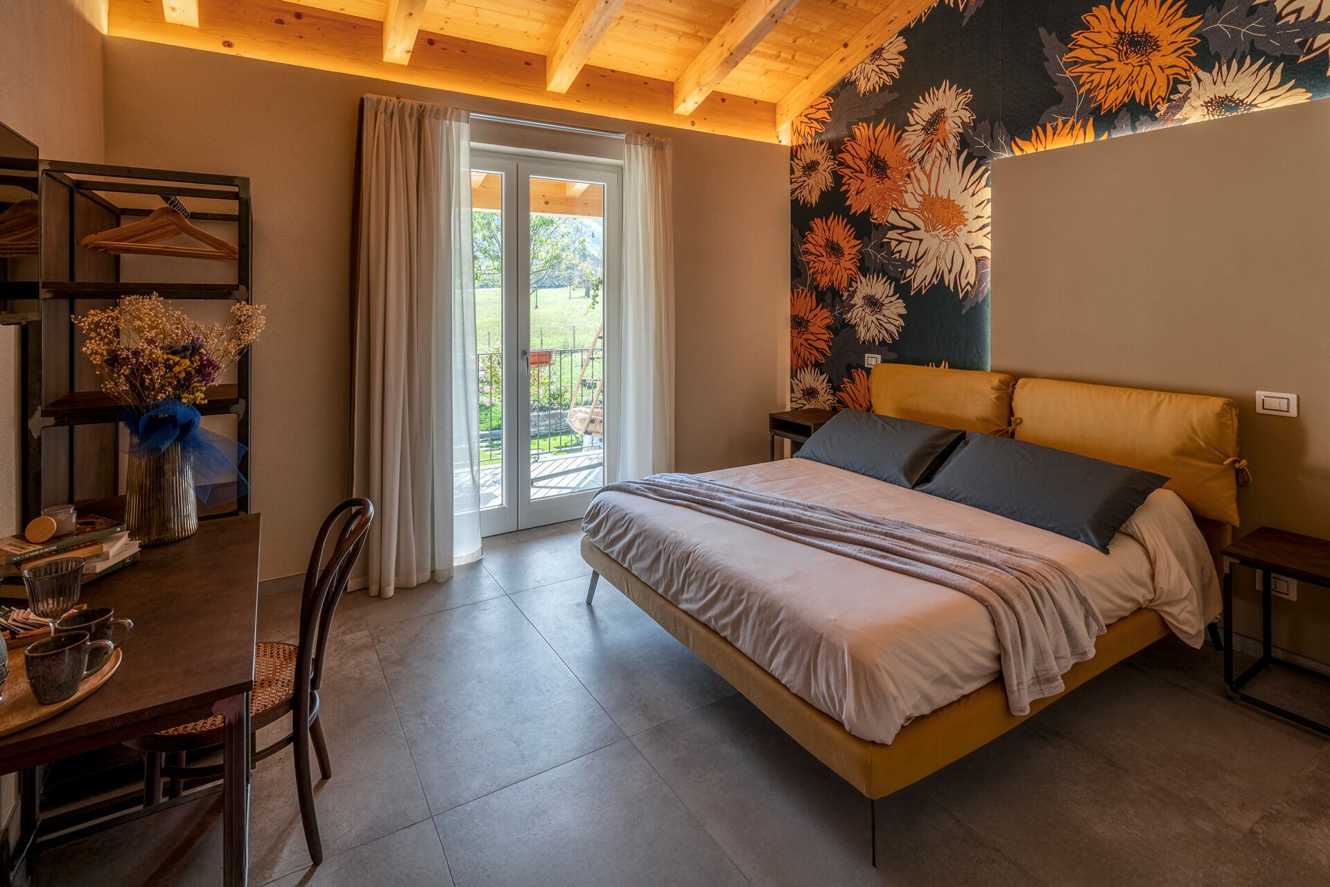 A bedroom with a bed, a desk with a vase of flowers, and a view through a glass door.