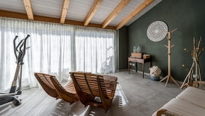 A room with a wooden ceiling, a large window with sheer curtains, a wooden bench, a bed, a small table with a vase, and a decorative wall piece.