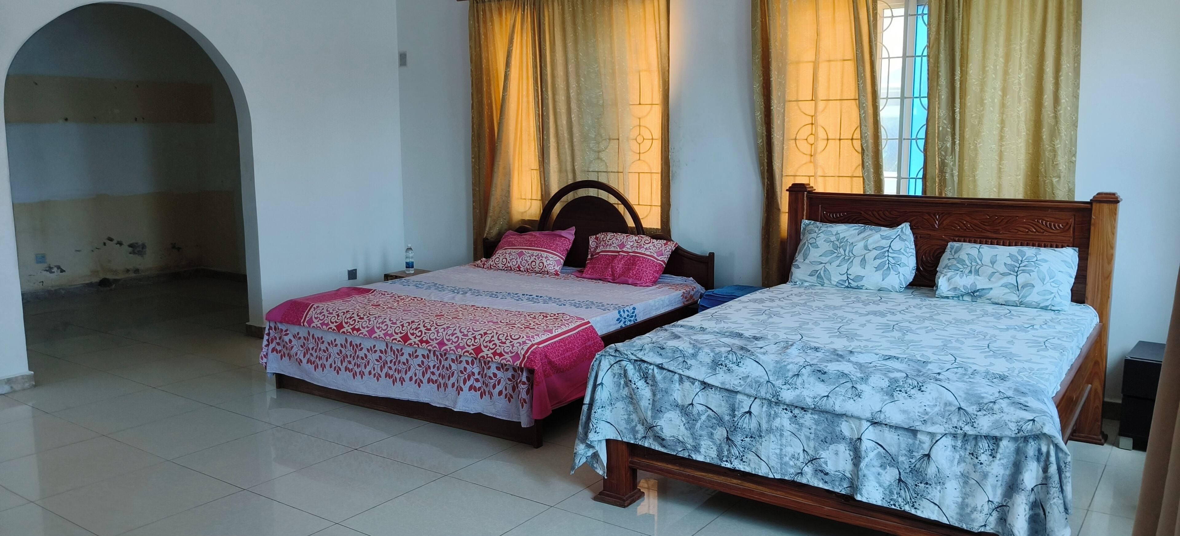 Habitación con dos camas, una con cubrecama floral y la otra con diseño de hojas. Hay una ventana con cortinas, piso de baldosas y una puerta que da a otra habitación.