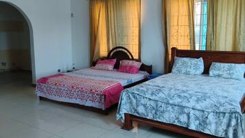 Habitación con dos camas, una con cubrecama floral y la otra con diseño de hojas. Hay una ventana con cortinas, piso de baldosas y una puerta que da a otra habitación.