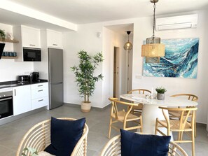 Eine moderne Küche mit weißen Schränken, einem Kühlschrank und einer Mikrowelle. Ein Essbereich mit einem runden Tisch und Korbstühlen. Ein großes Gemälde hängt an der Wand, und eine Topfpflanze steht in der Ecke.