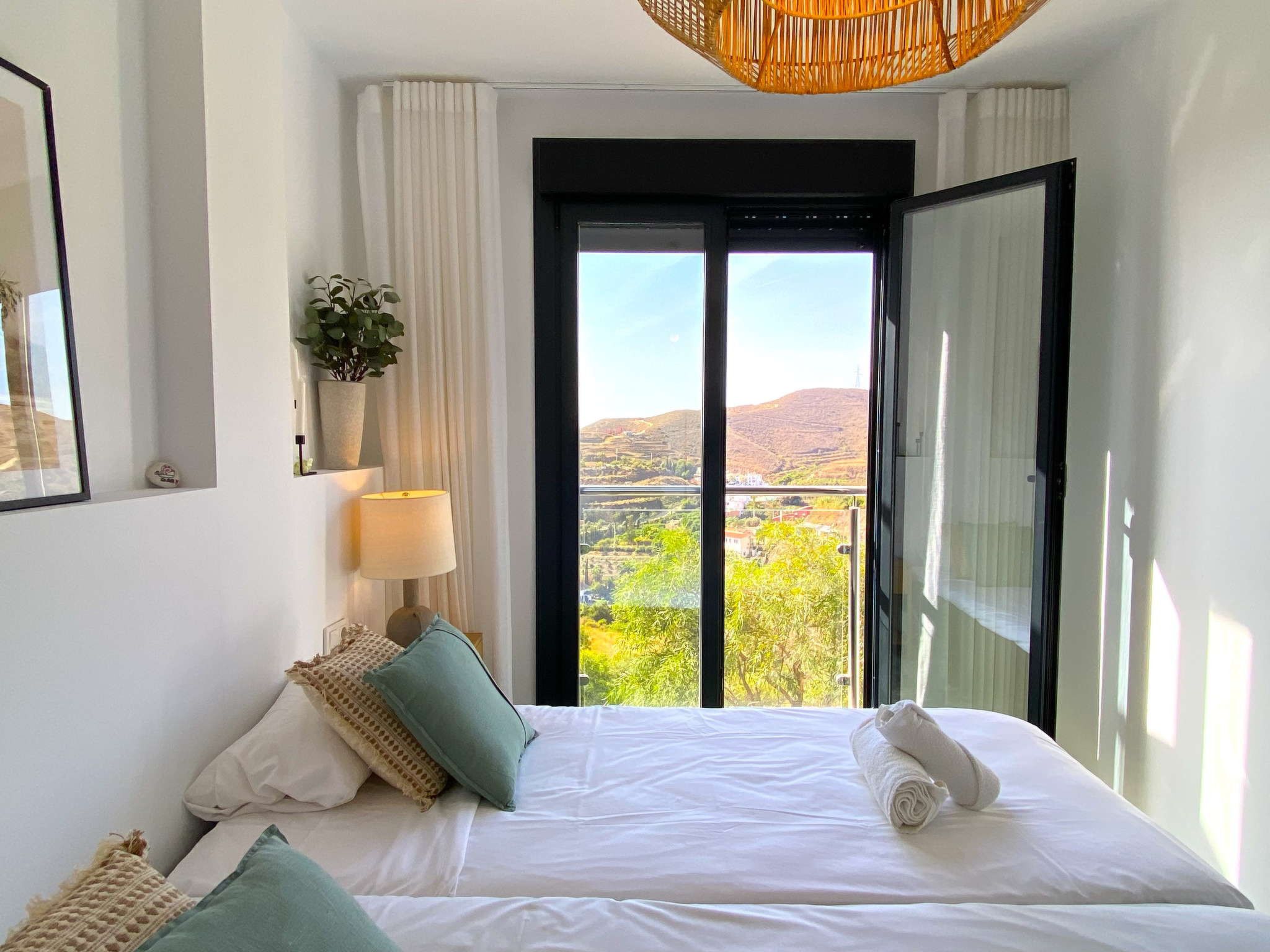 A hotel room with a bed, a bedside lamp, a potted plant, and a view of a landscape through a large window.