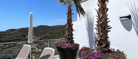 A terrace with two lounge chairs, a white wall, and a view of a hilly landscape.