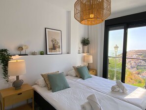 Ein Hotelzimmer mit einem großen Bett, zwei Nachttischen, einer Pendelleuchte, einem gerahmten Bild und Blick auf eine Stadtansicht durch ein großes Fenster.