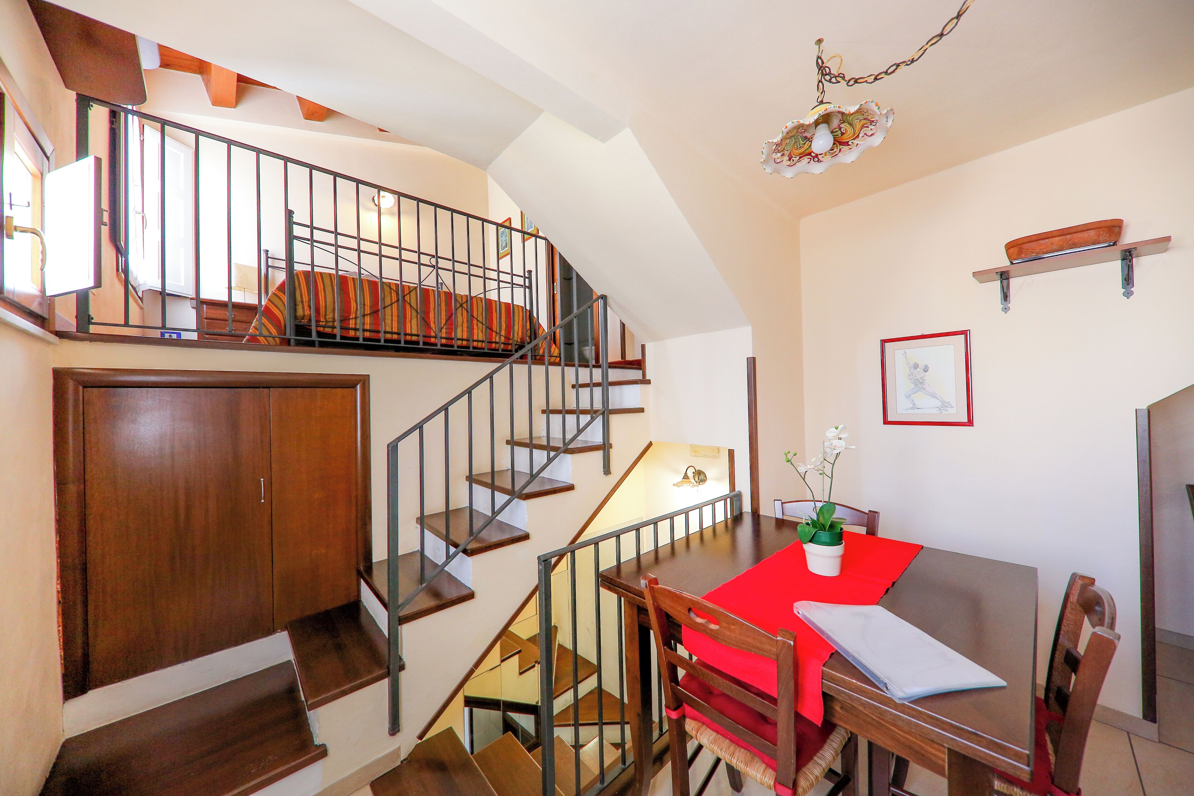 A staircase with a wooden finish, a dining area with a red tablecloth, and a wall-mounted shelf with a framed picture.