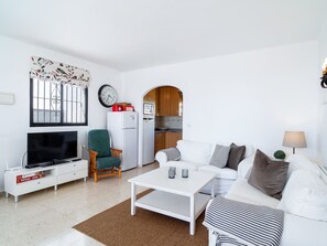 A living room with a television, a sofa, a coffee table, a clock, and a kitchen visible through an archway. - Los Naranjos 15B Capistrano Village by Casasol by Interhome (Nerja)