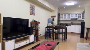 Ein Wohnzimmer mit Flachbildfernseher, Couchtisch, Essbereich mit Tisch und Stühlen sowie eine Küche mit weißen Schränken und Geräten.