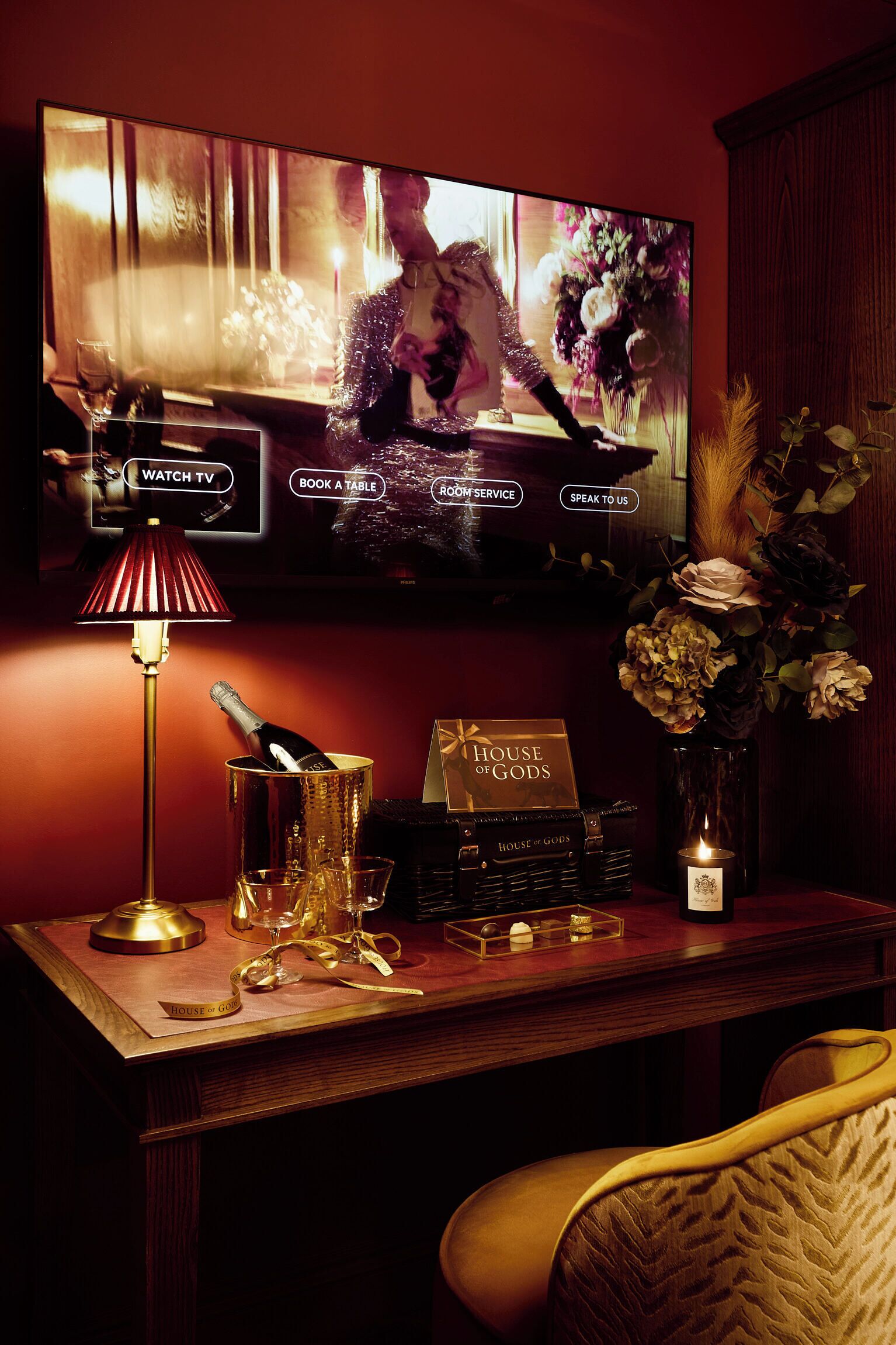 Un comptoir d’accueil d’hôtel avec un téléviseur affichant un menu, une lampe, une bouteille de champagne et un vase rempli de fleurs.