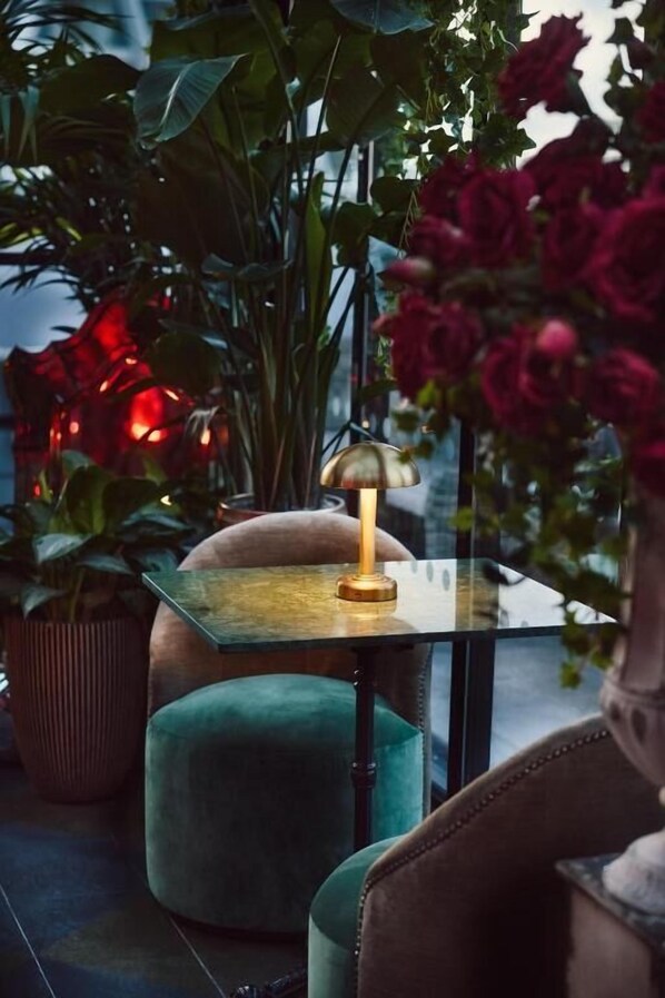 A table with a lamp, surrounded by potted plants and a green ottoman.