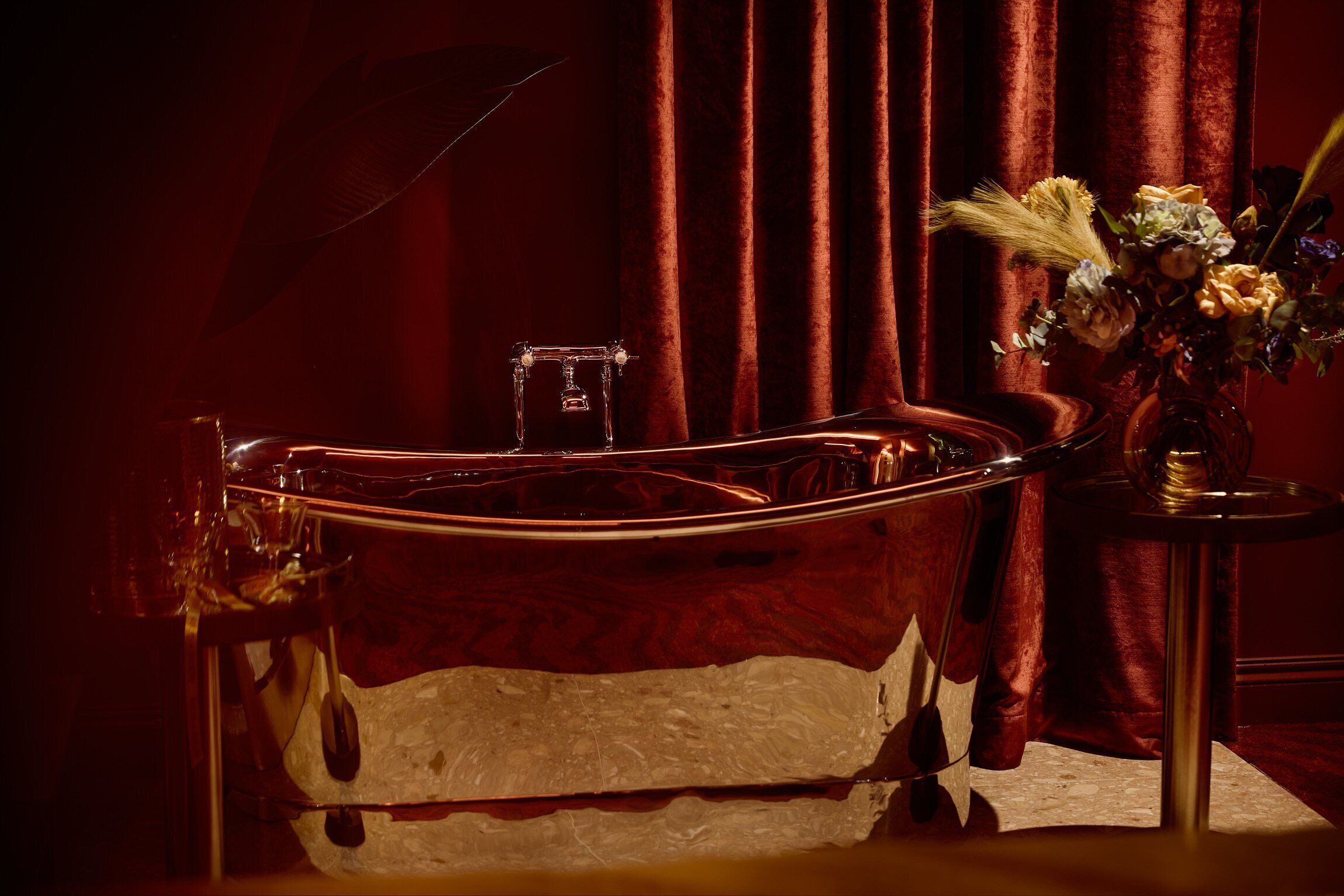 A bathtub with a glass enclosure, a small glass container, and a vase with flowers on a side table.