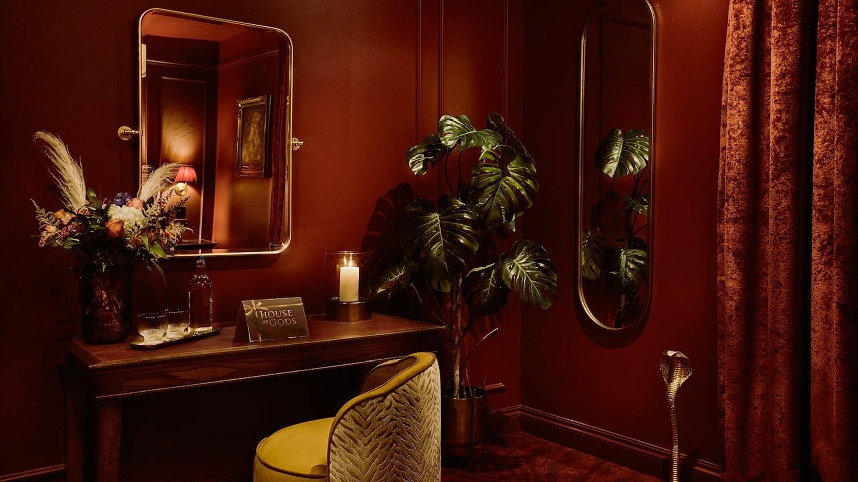 A dimly lit room with a wooden vanity, a mirror, a potted plant, a candle, and a sign that reads 'HOUSE OF GODS'.