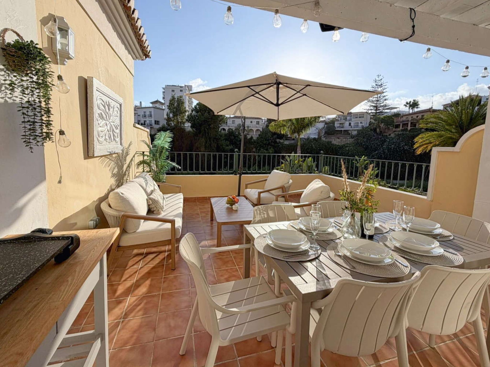 A sunlit outdoor dining area with a table set for four, surrounded by white chairs, a beige umbrella, and a view of buildings in the background.