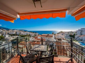 Ein Balkon mit Tisch und Stühlen, der Blick auf eine Küstenstadt mit Gebäuden und dem Meer freigibt.