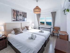 Ein Hotelzimmer mit zwei Betten, einem Schreibtisch, einem Stuhl, einem Fenster mit Meerblick und einer Pendelleuchte.