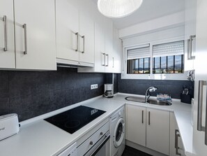 Uma cozinha moderna com armários brancos, forno embutido, máquina de lavar e janela com vista para a cidade.