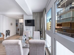 Ein modernes Wohnzimmer mit Flachbildfernseher, einem Essbereich mit Glastisch und Blick auf ein Gebäude mit Balkonen.