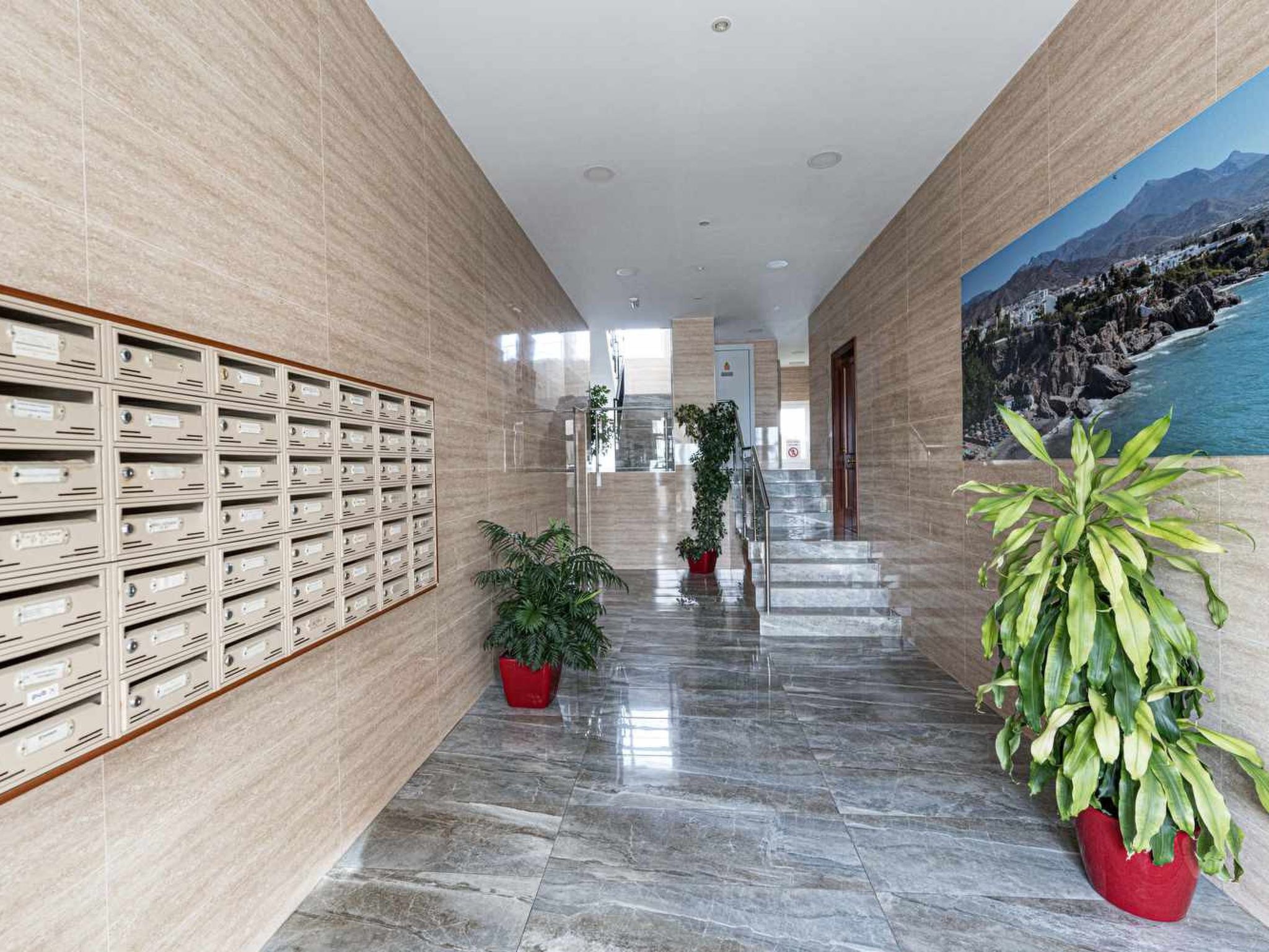 Un couloir avec une rangée de boîtes aux lettres sur le côté gauche, des plantes en pot et, sur le côté droit, un grand tableau représentant un paysage côtier.