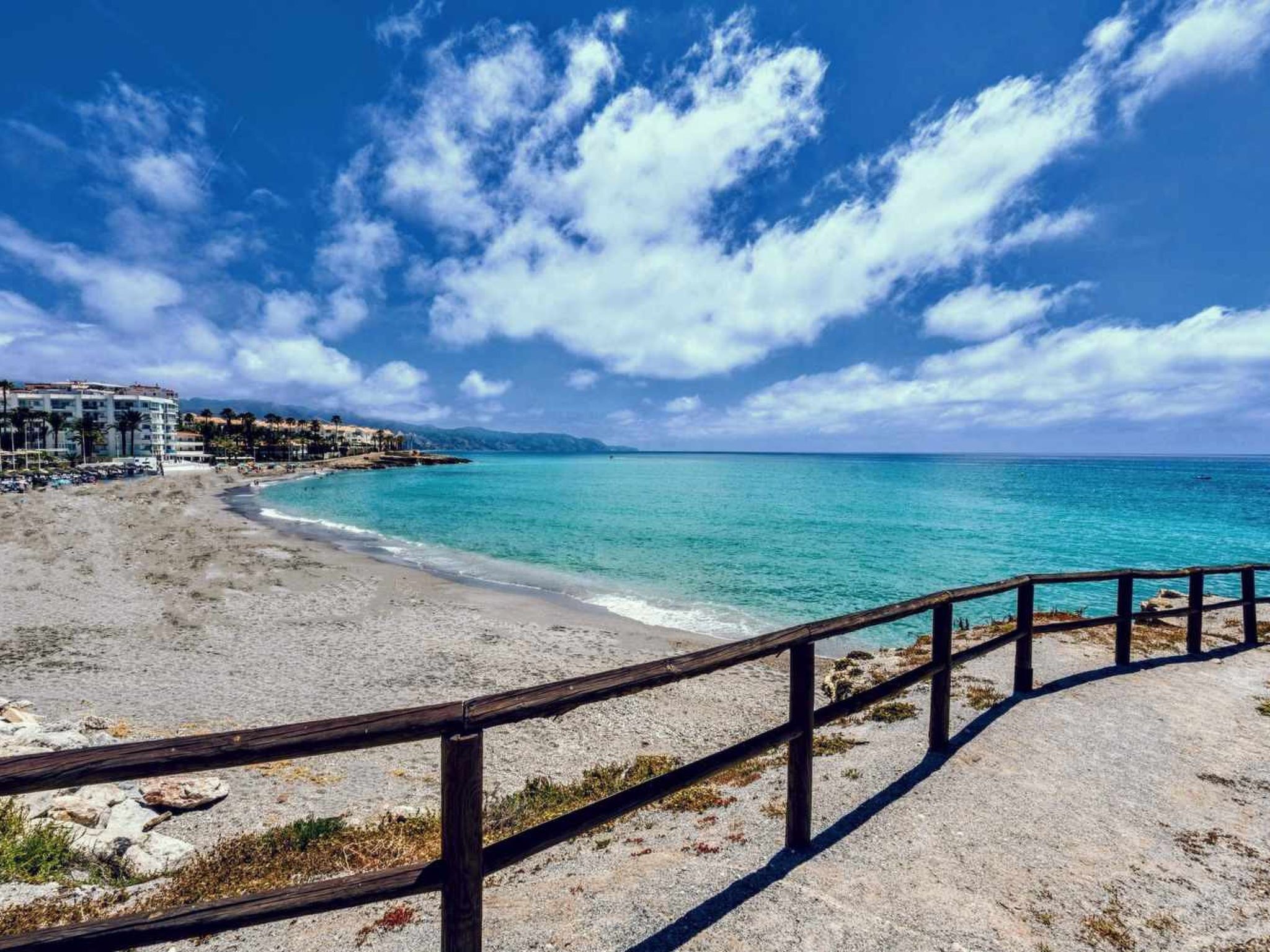 Ein Strand mit Holzlattenzaun, klarem türkisblauem Wasser und einem Gebäude im Hintergrund.