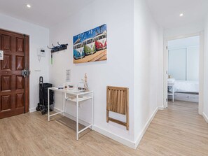 Um quarto com porta de madeira, uma mesa branca com uma cadeira, um radiador e uma cama com colcha branca.