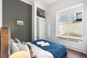 A bedroom with a large window, a bed with a blue blanket, and a painting on the wall. - Seaview Haven By My Getaways (Brighton)