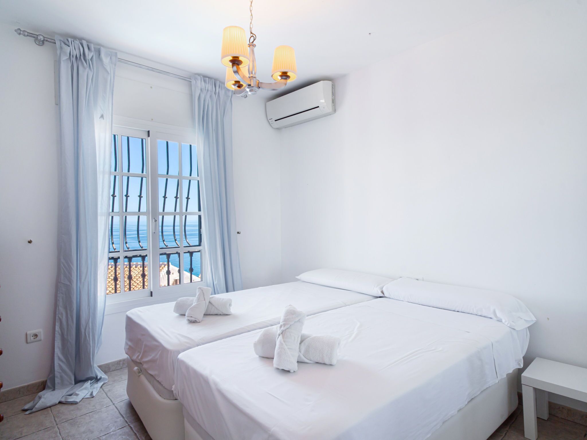 Een hotelkamer met een bed, nachtkastje, airconditioning en uitzicht op de zee door een raam met gordijnen.