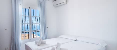 A hotel room with a bed, a bedside table, an air conditioning unit, and a view of the sea through a window with curtains.