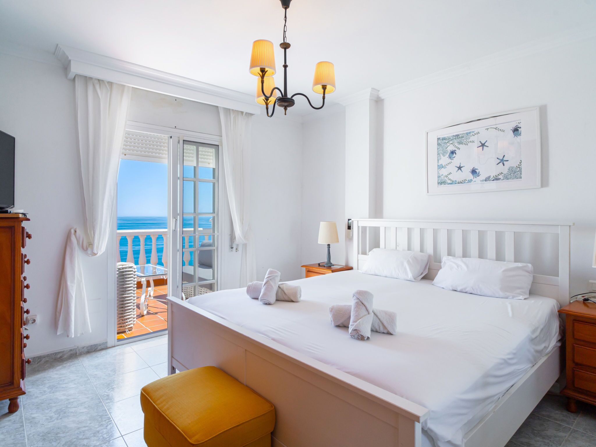 Una habitación de hotel con cama, mesita de noche, lámpara, un cuadro y vista al mar a través de una puerta a un balcón.