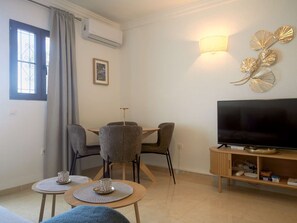A living room with a TV, a dining table with chairs, a sofa, and a wall-mounted light fixture. - Casa Sofia Capistrano Village Pueblo 110B by Interhome (Nerja)