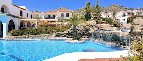A swimming pool with a waterfall feature, surrounded by white buildings with red-tiled roofs, and a mountainous backdrop.
