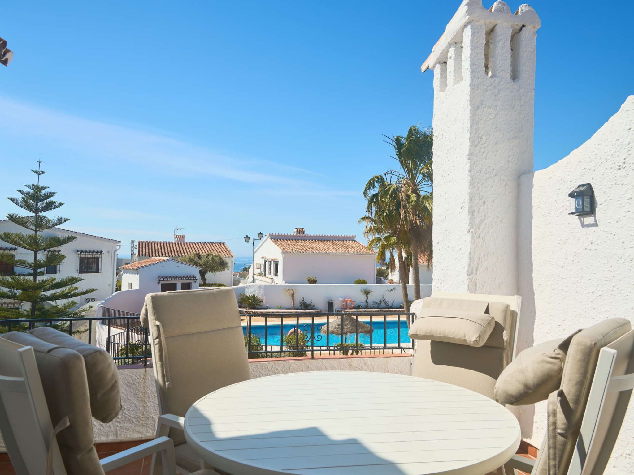 A poolside area with a round table and chairs, a white building with a tower, and a clear blue sky.