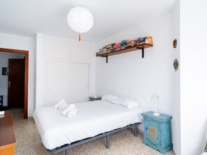 Ein Schlafzimmer mit einem Bett, weißen Wänden, einer weißen Tür, einer weißen Deckenleuchte, einem kleinen Nachttisch mit Lampe und einem Bücherregal.