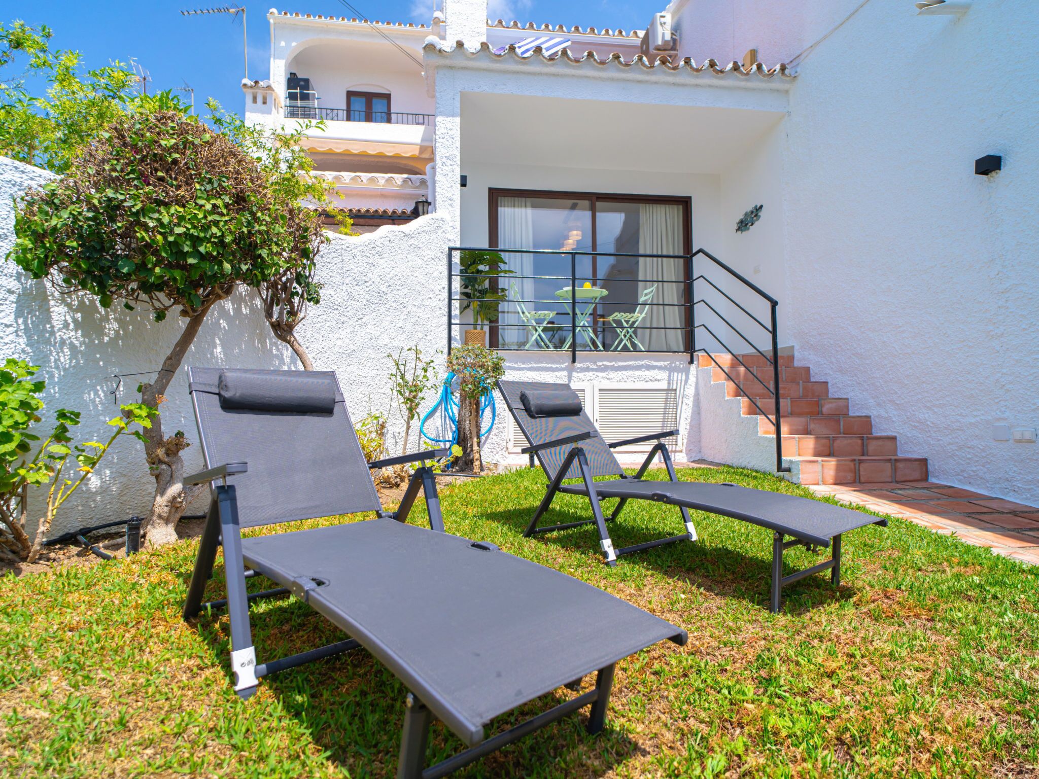 Eine Terrasse mit zwei Liegestühlen und einem kleinen Tisch, ein Baum und ein Gebäude mit Balkon.