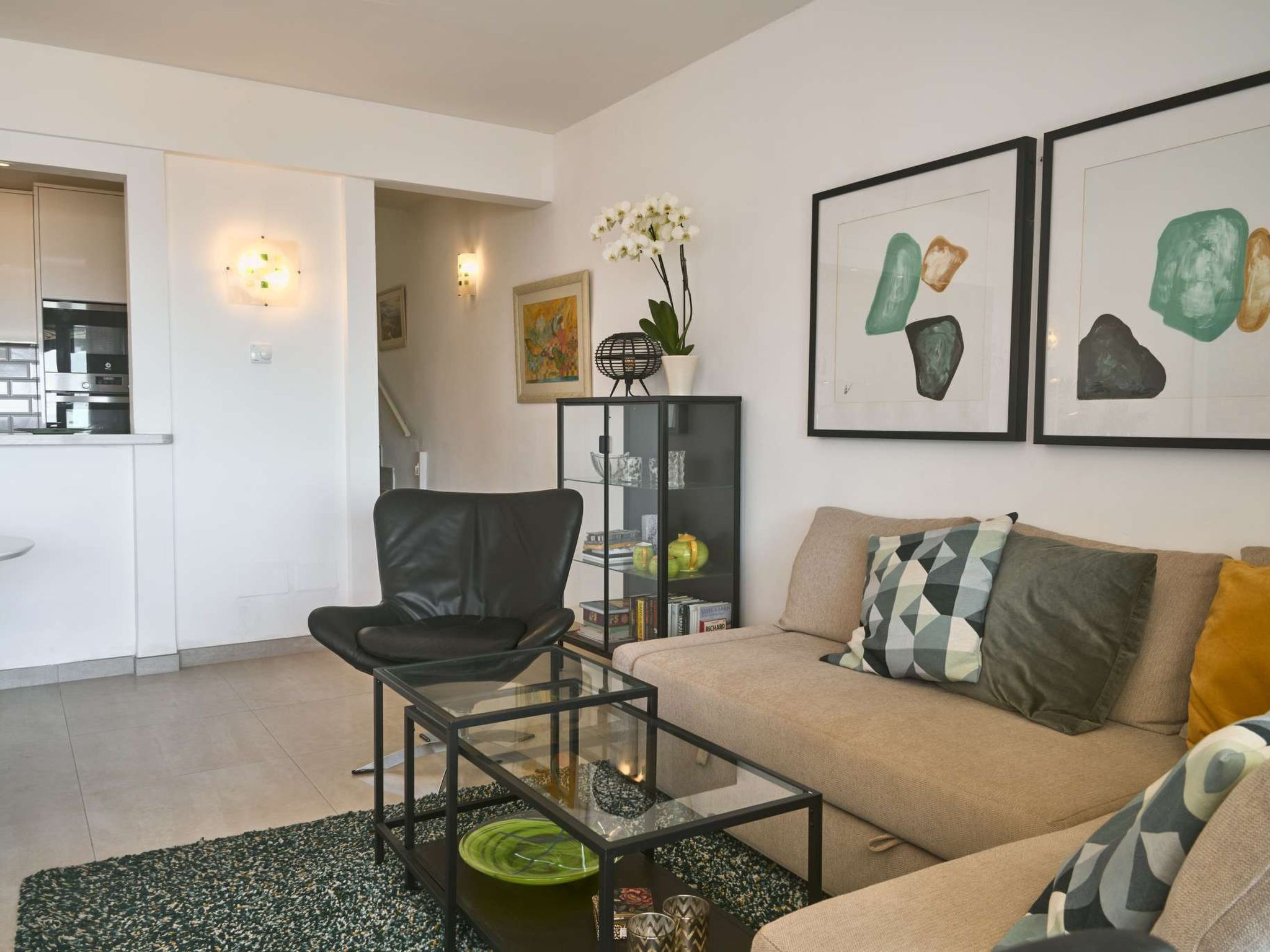 Un salon moderne avec un fauteuil en cuir, une table basse en verre, un canapé agrémenté de coussins à motifs et des œuvres d’art accrochées au mur.