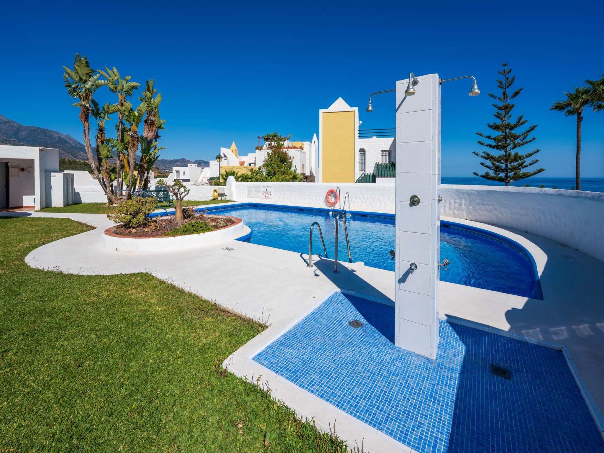 A swimming pool with a clear blue tile design, surrounded by a white pool deck, with a view of the sea and buildings in the background.