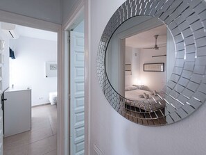 Ein Flur mit einem runden Spiegel an der Wand, ein Bett mit Kopfteil im angrenzenden Raum und eine weiße Tür zu einem weiteren Zimmer.