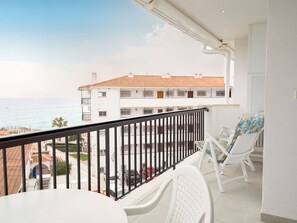 Ein Balkon mit Meerblick, weiße Möbel und ein Gebäude mit einem Ziegeldach im Hintergrund.