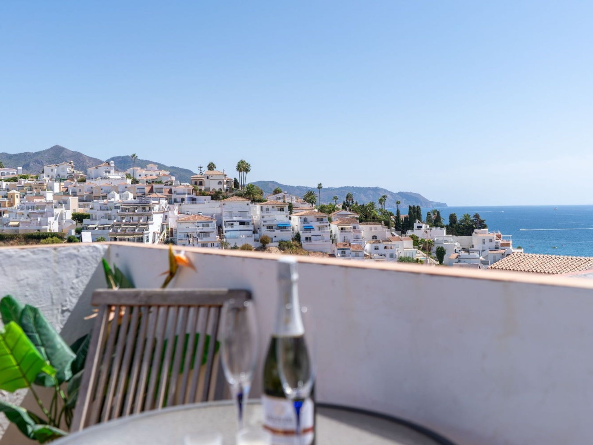 A terrace with a table, two glasses, and a bottle, overlooking a coastal town with white buildings and a clear sky.