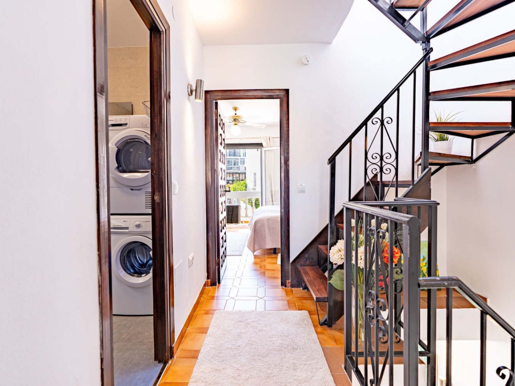 A hallway with a washing machine, a staircase with a railing, and a view into a room with a bed.