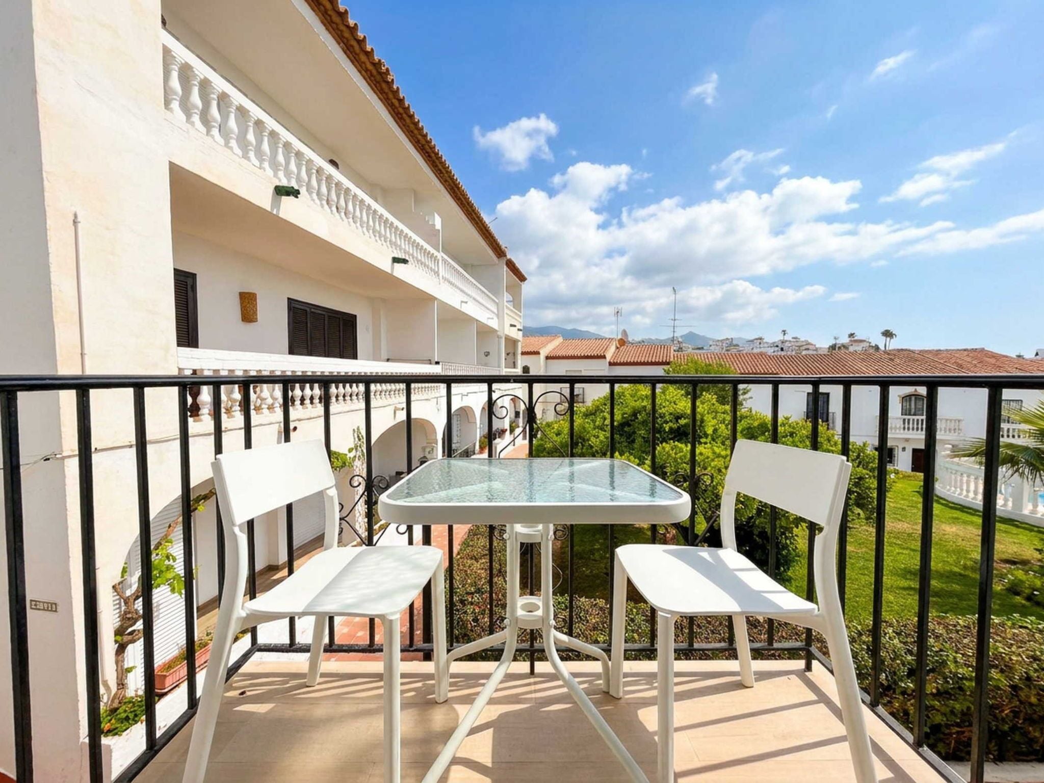 A balcony with a glass table and two white chairs, overlooking a residential area with terracotta roofs.
