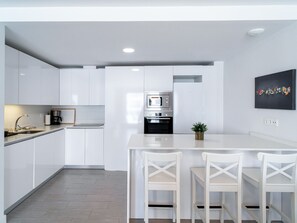 A modern kitchen with white cabinetry, a central island with chairs, a built-in oven, and a coffee machine. - Duna Beach Torrox Costa by Casasol by Interhome (Torrox Costa)